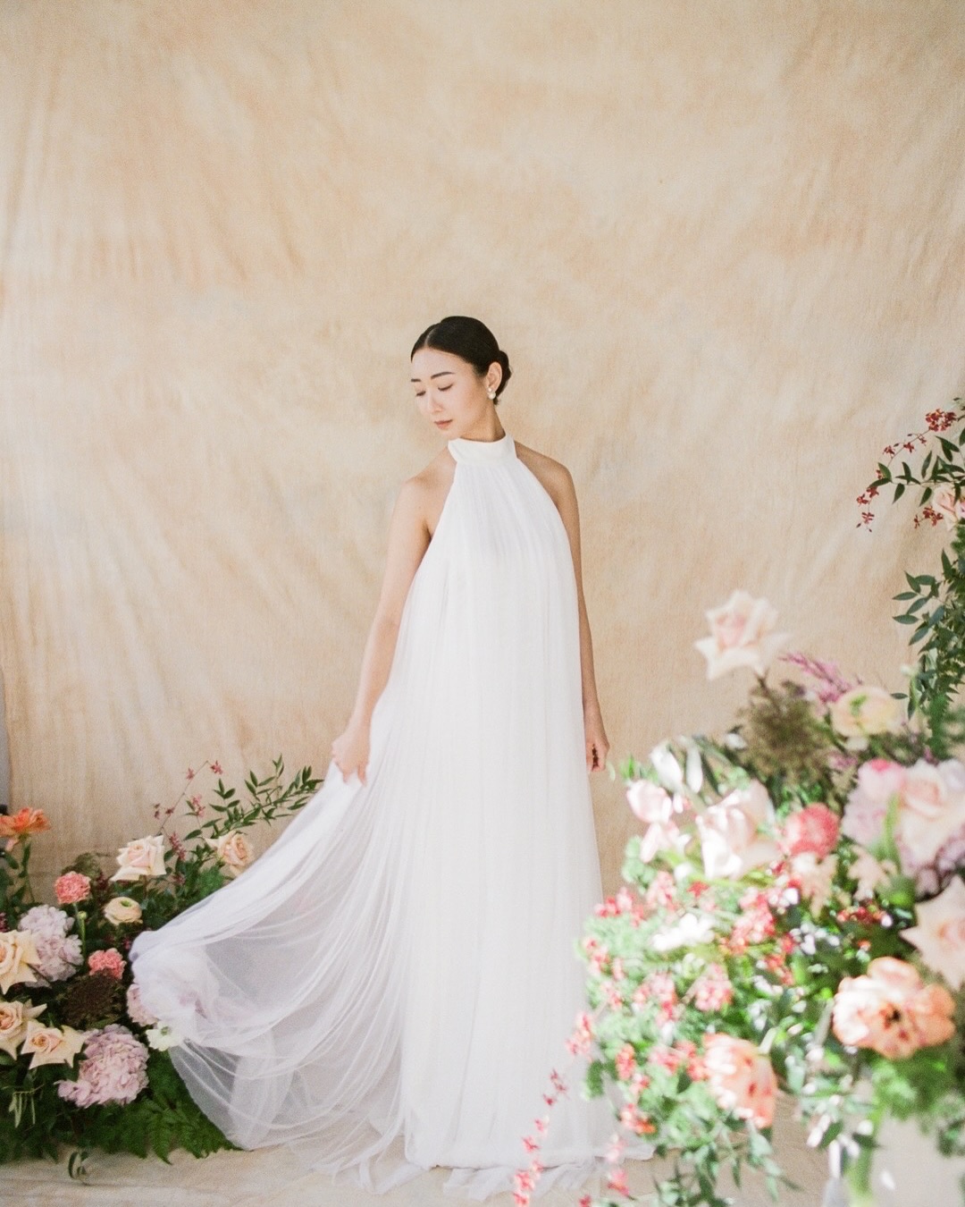 A bride in a flowing high-neck gown stands beside low arrangements of pink and cream flowers