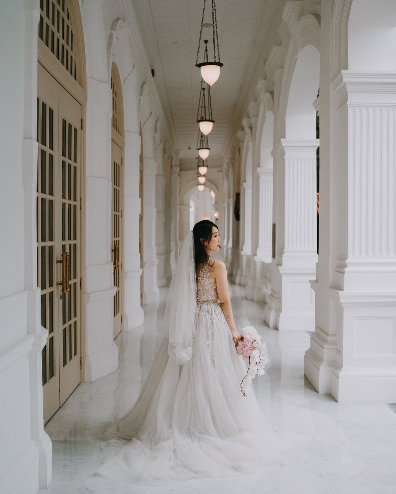 A bride with a long veil and bouquet stands in a white heritage colonnade with marble floors