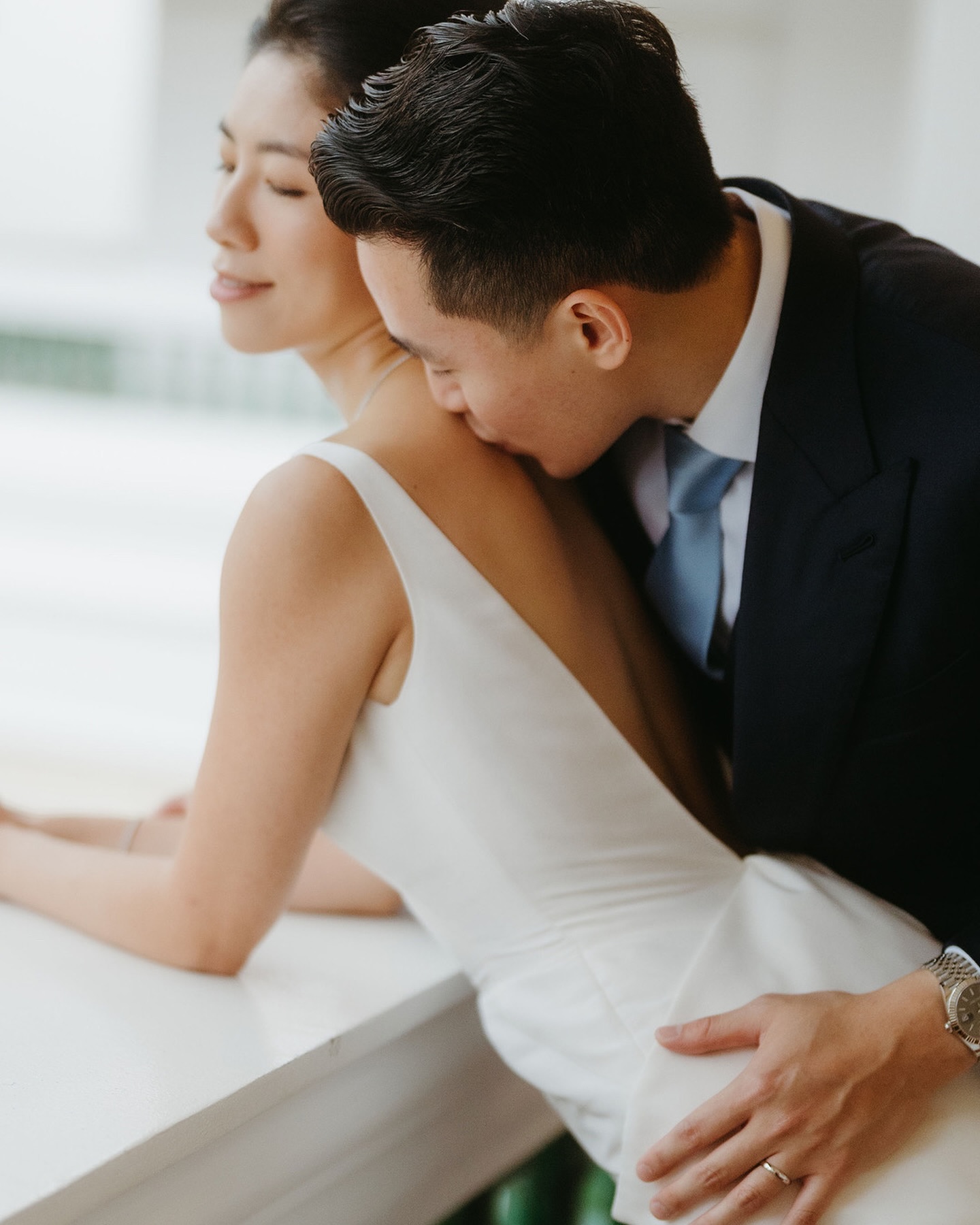 A groom leans in to kiss the bride's shoulder as she looks out a window, both in quiet close-up