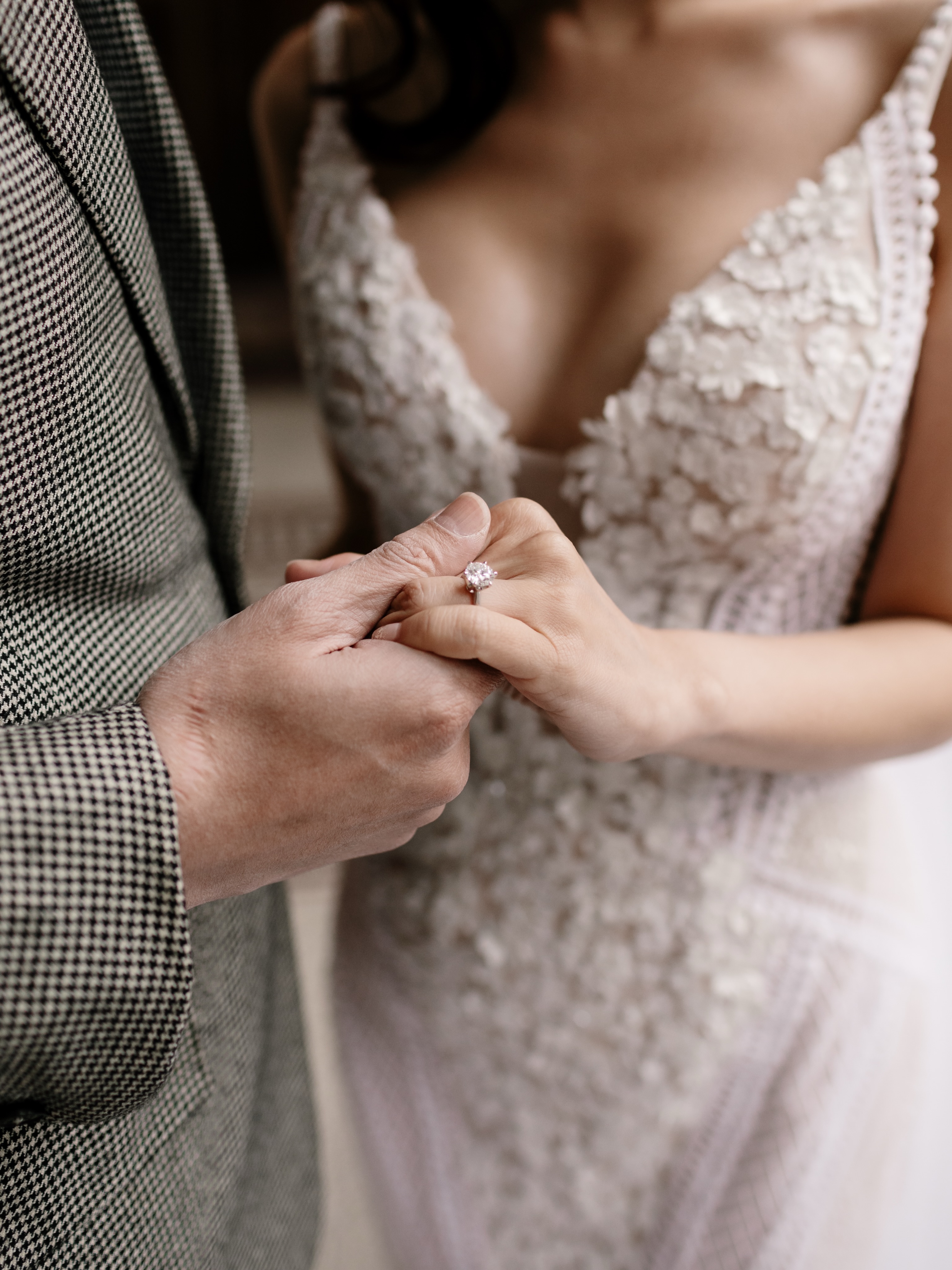 Close-up of a bride's hand with a solitaire diamond ring resting in another hand, lace gown behind